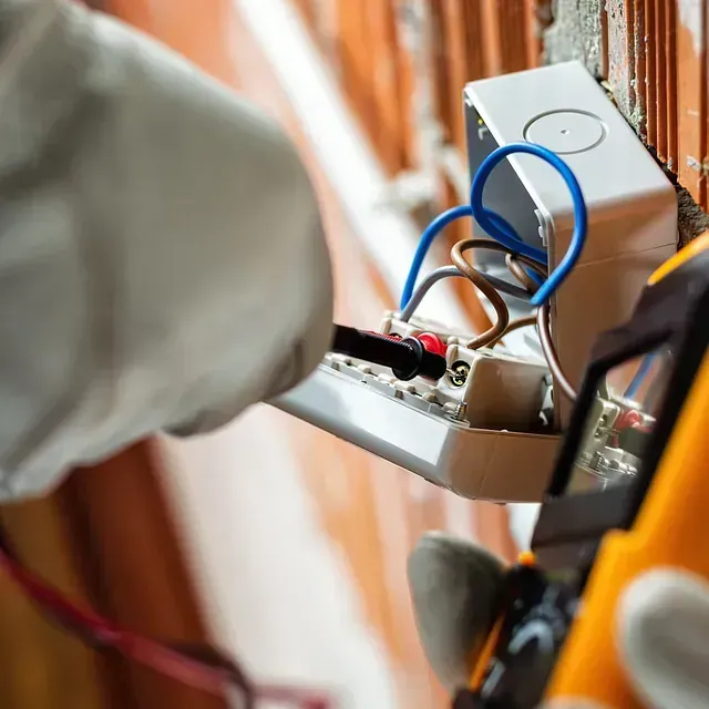 Electricista prueba cables en una caja eléctrica montada en la pared con un multímetro; manos enguantadas visibles.
