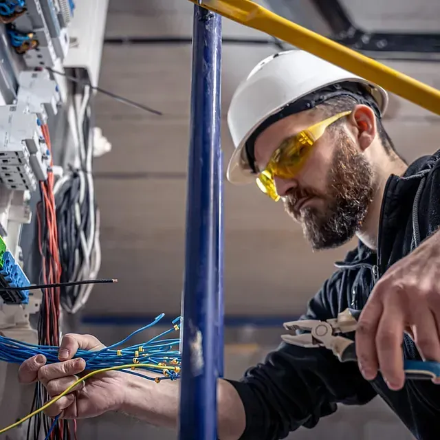 Un electricista con casco blanco y gafas de seguridad trabaja en el cableado eléctrico.