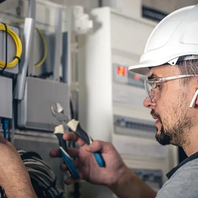 Electricista con casco blanco y gafas de seguridad trabajando en cableado eléctrico.