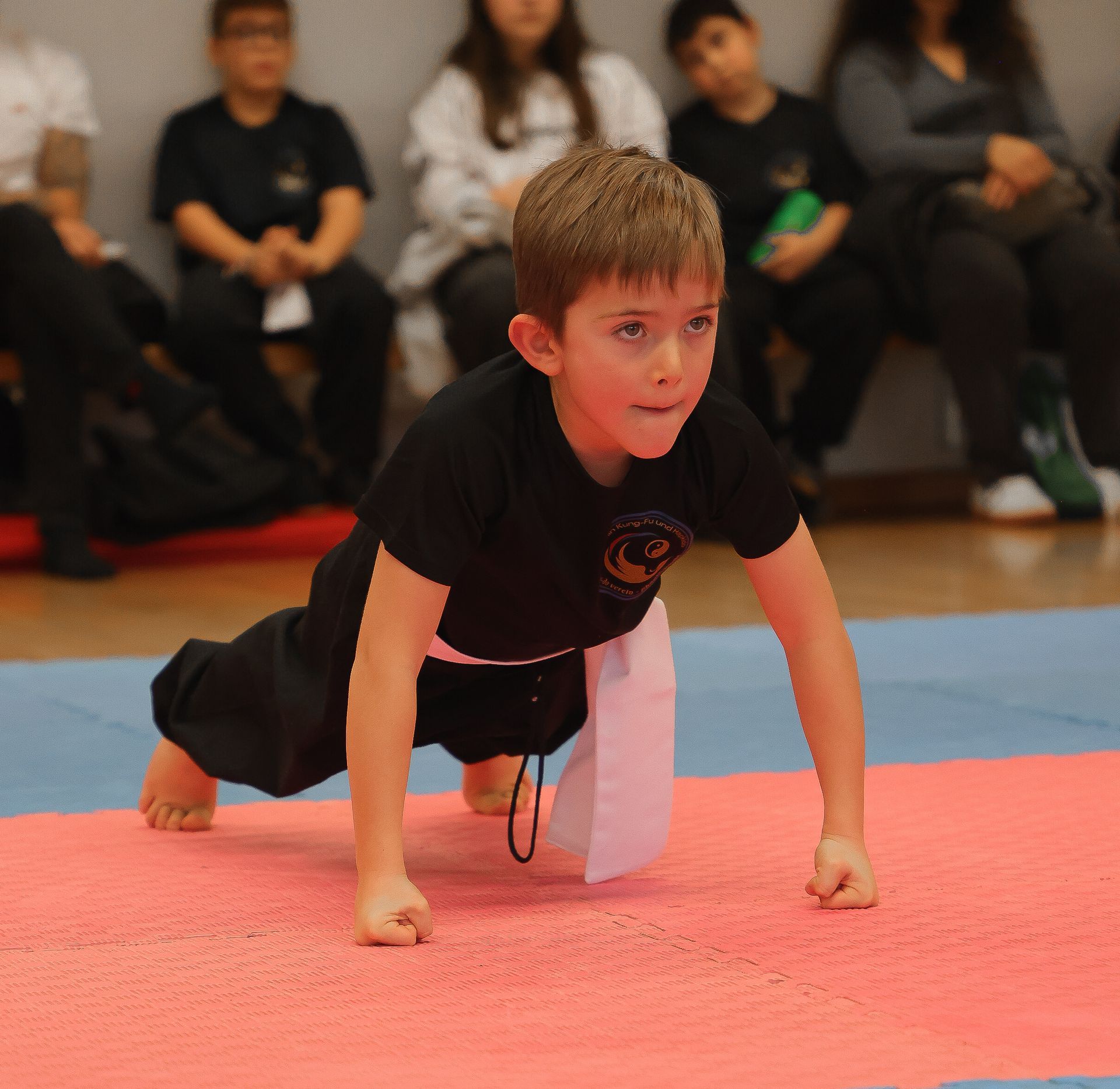Ein Junge in schwarzer Kampfsportuniform macht Liegestütze auf einer Matte; im Hintergrund sitzen Zuschauer.
