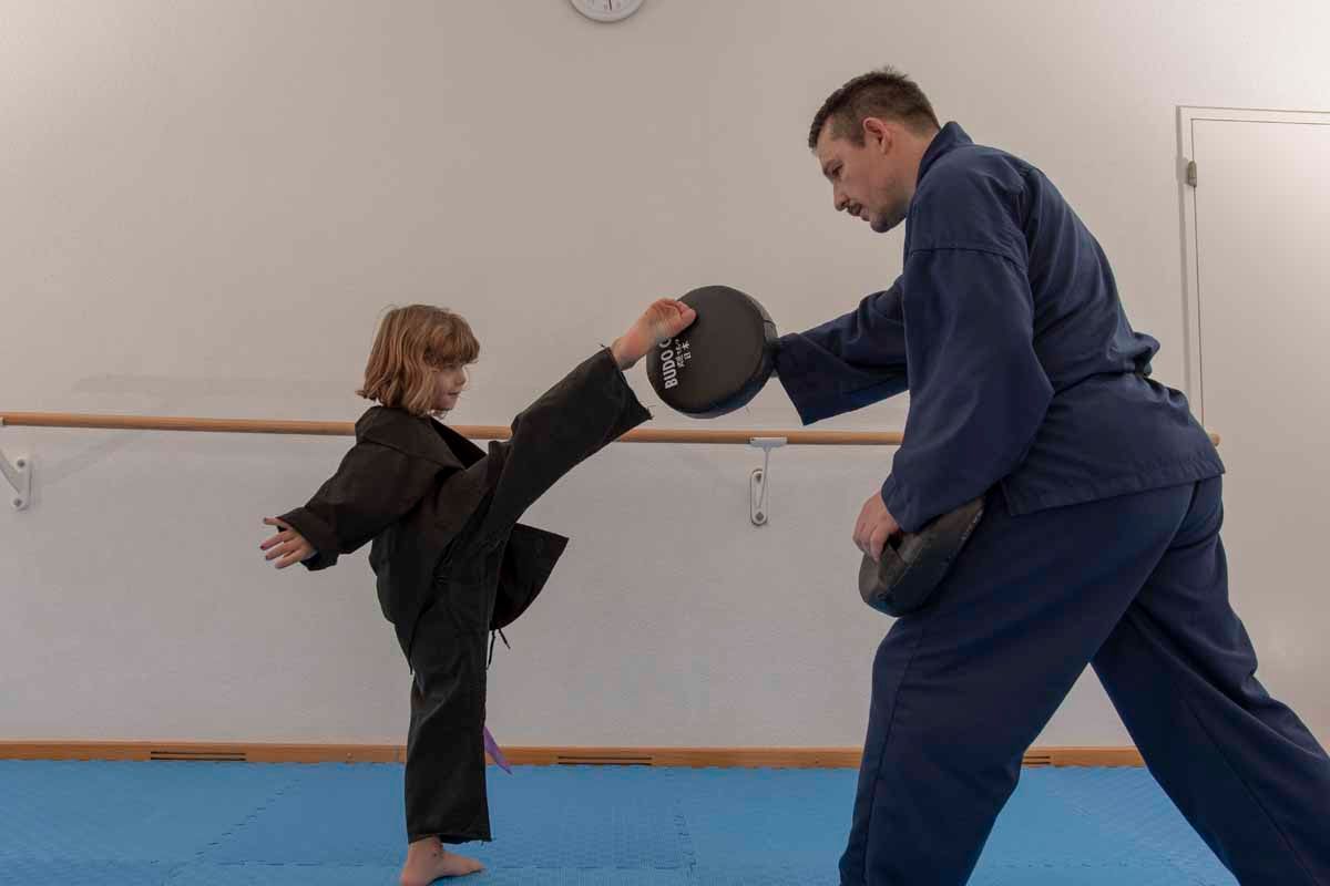 Ein Kind tritt in einem Kampfsportkurs gegen einen von einem Erwachsenen gehaltenen Schlagpolster.