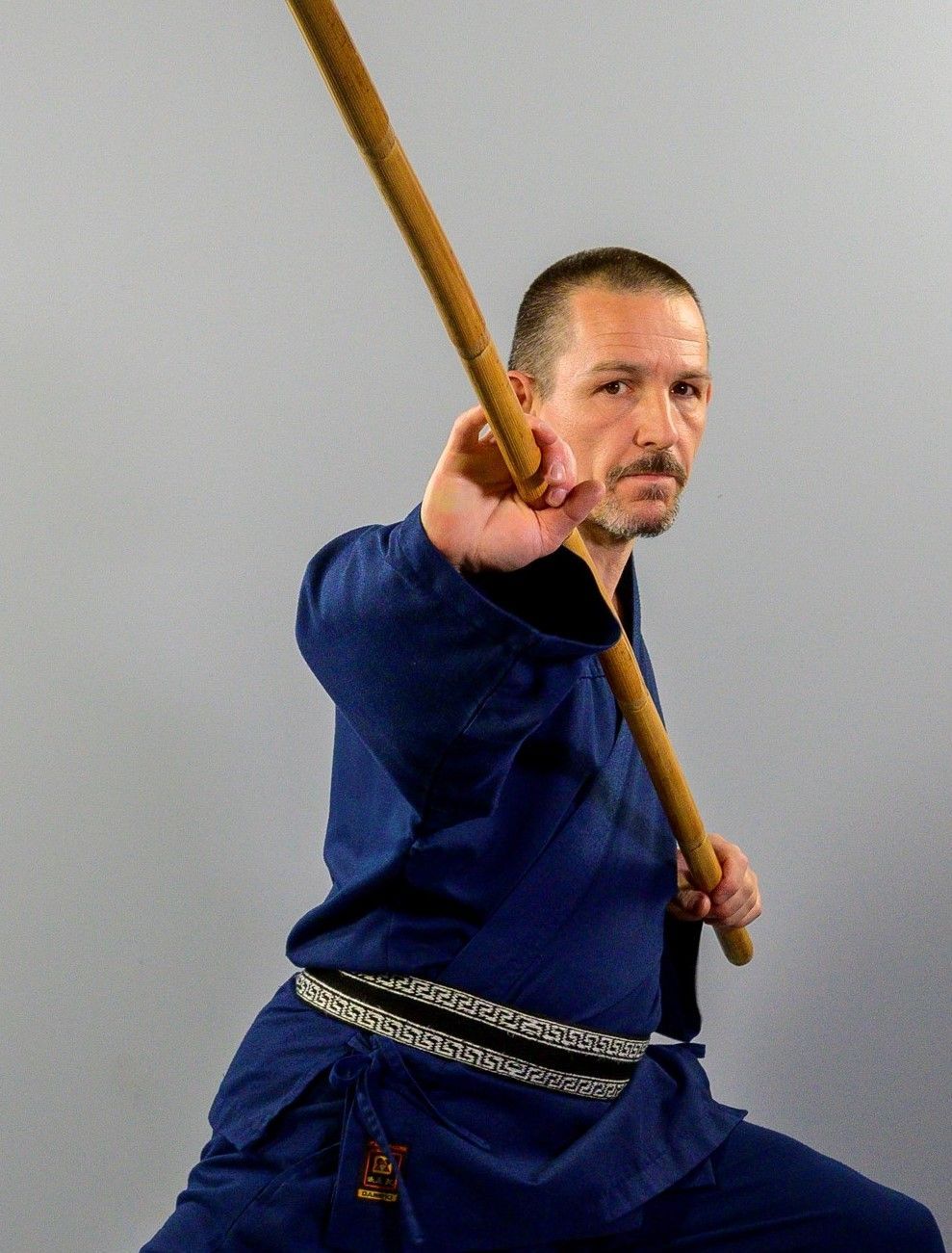 Mann in blauer Kampfsportuniform mit Stab, der einen Schlag ausführt, grauer Hintergrund.
