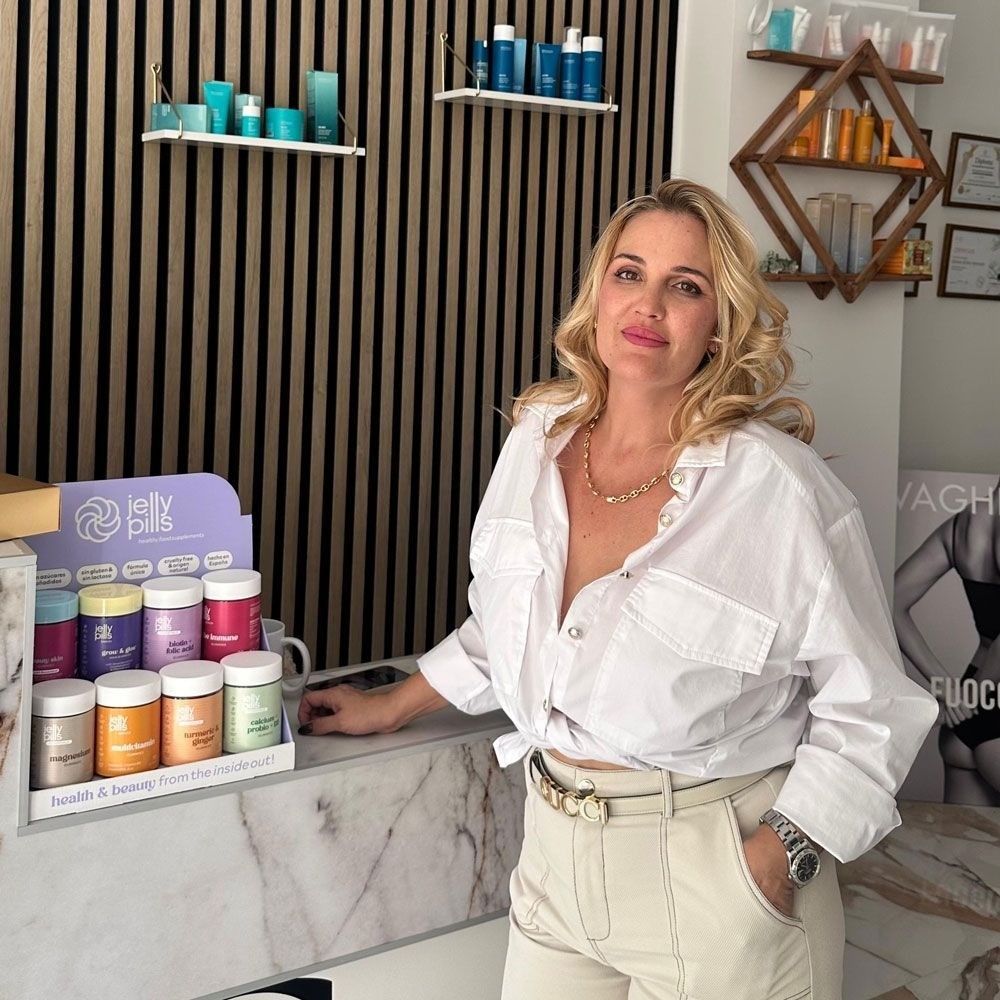 Mujer con una blusa blanca de pie detrás de un mostrador de mármol con frascos de colores 