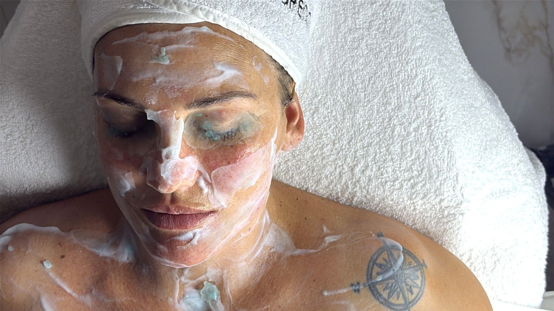 Persona tumbada boca arriba con una mascarilla facial blanca y espuma, relajándose sobre una toalla de spa