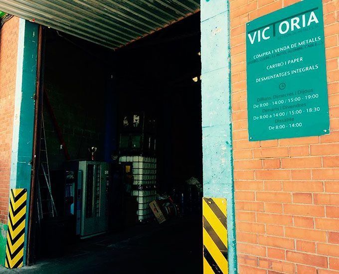 Entrada a un centro de reciclaje, pared de ladrillo con un letrero verde. El letrero dice "Victoria" y el horario de apertura.