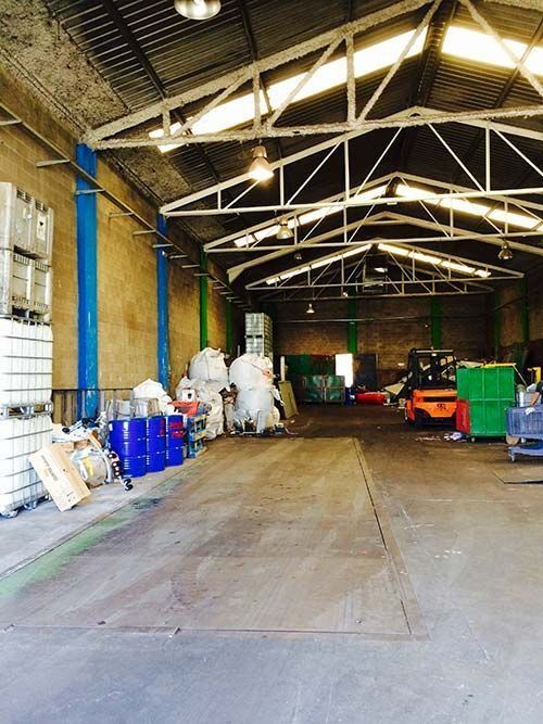 Interior de almacén con barriles azules, una carretilla elevadora y varios contenedores industriales.