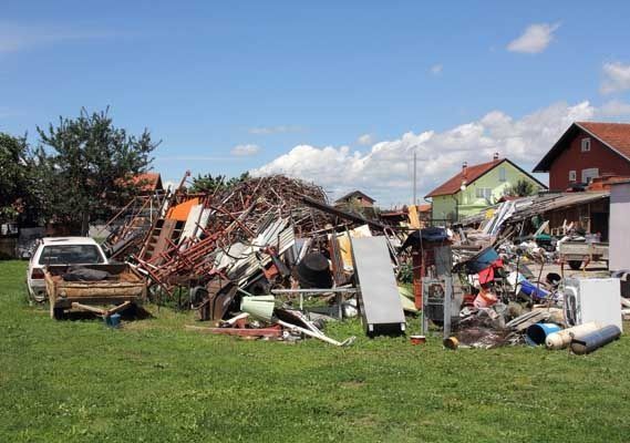 Pila de chatarra y escombros en un patio, con un coche blanco y casas al fondo.