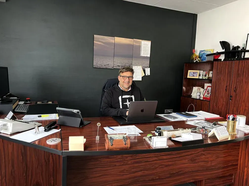 Un hombre sentado en un escritorio de oficina desordenado con una computadora portátil, estantes y papeles detrás.