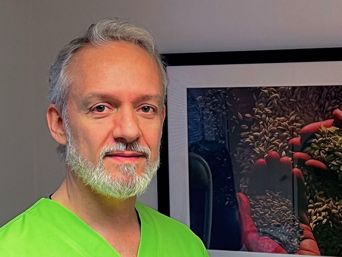 Un hombre con barba y una blusa verde está parado frente a un cuadro.