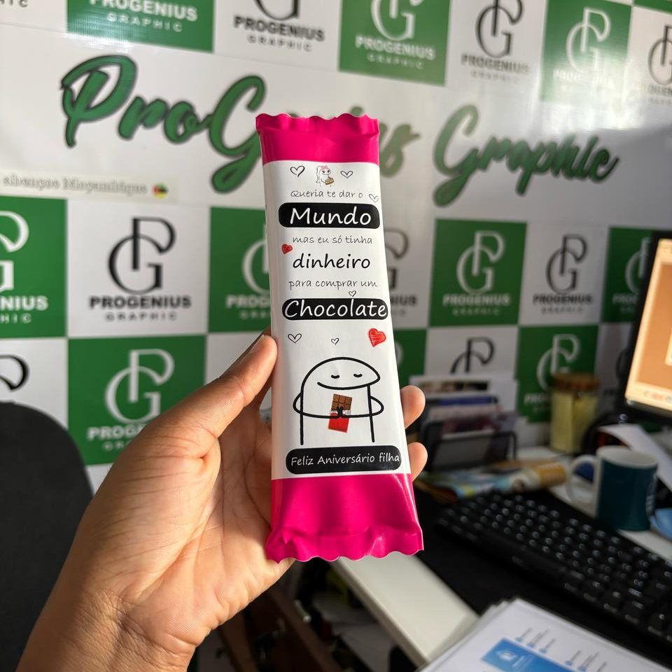Mão segurando uma barra de chocolate embrulhada em rosa com a inscrição “Mundo de dinheiro” em um ambiente de escritório.
