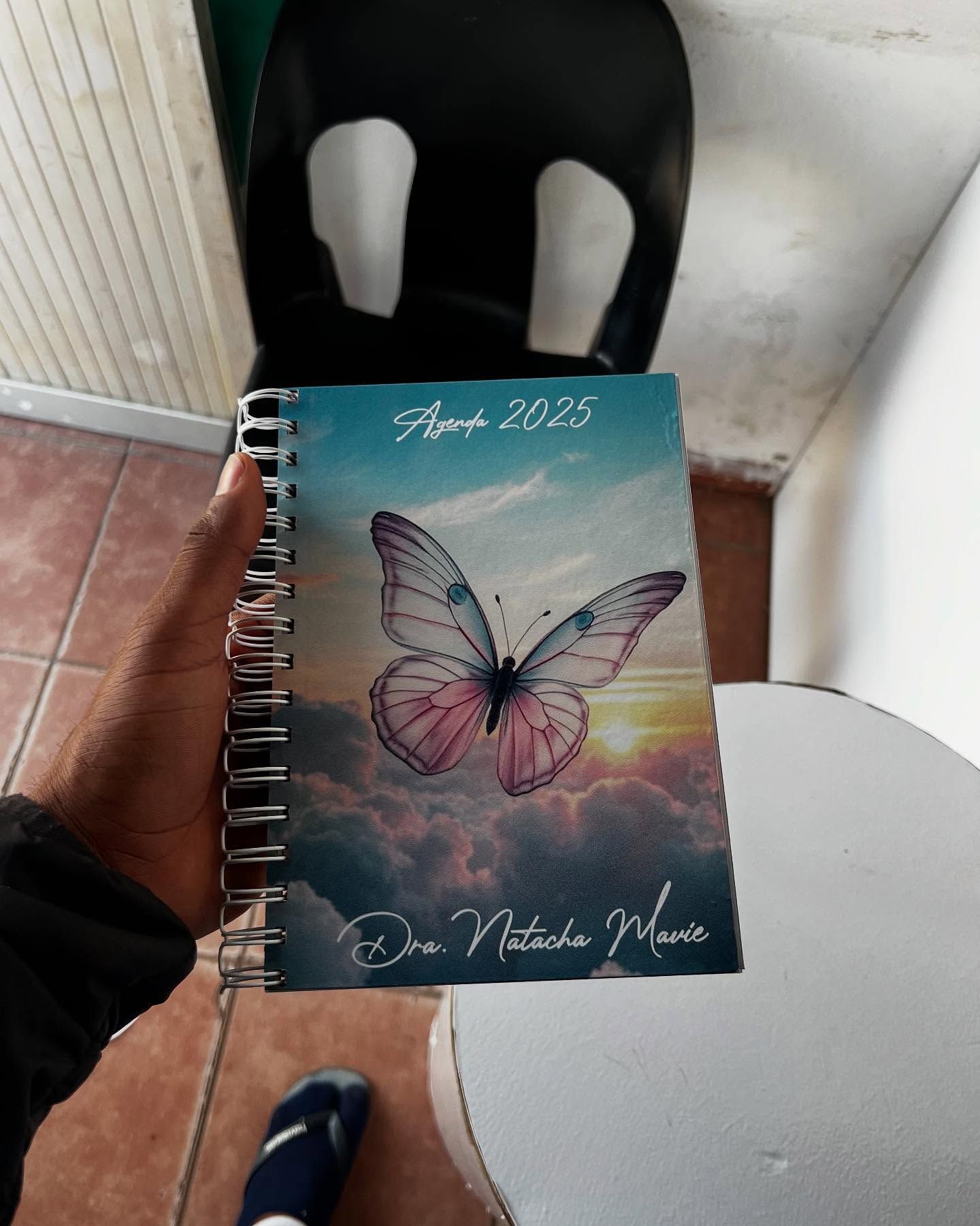 Agenda com borboleta rosa no colo de uma pessoa, em ambiente interno, com capa azul com céu e nuvens e a inscrição "2023".