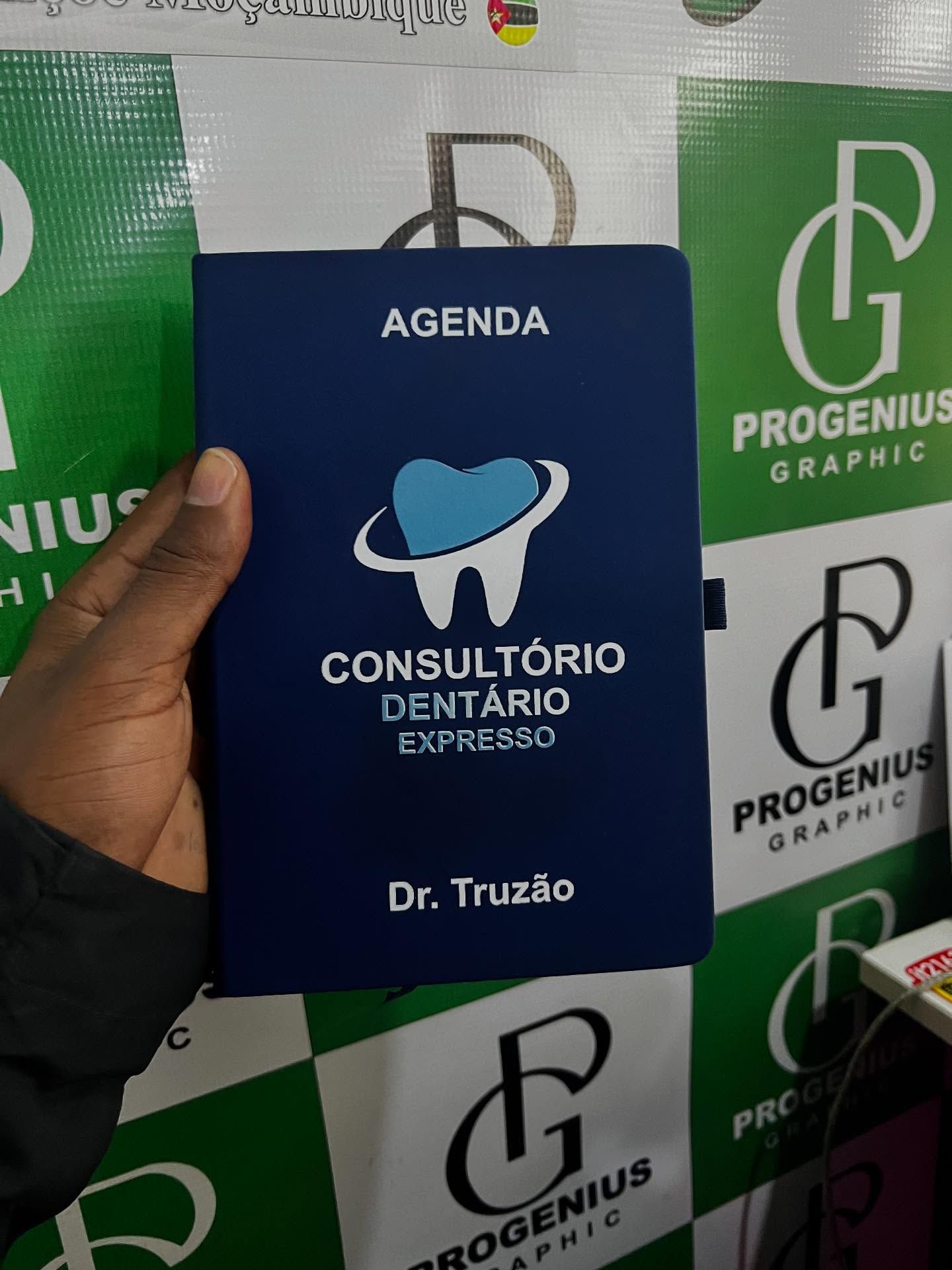 Mão segurando uma agenda de consultas odontológicas azul com o logotipo de um dente e a inscrição “Dr. Truzão” na capa.