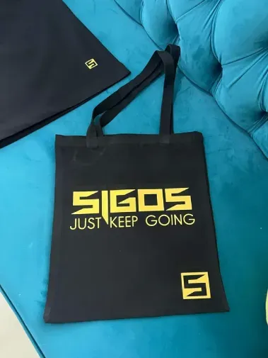Bolsa preta da SIGOS com a frase “JUST KEEP GOING” em amarelo sobre um sofá azul-petróleo.
