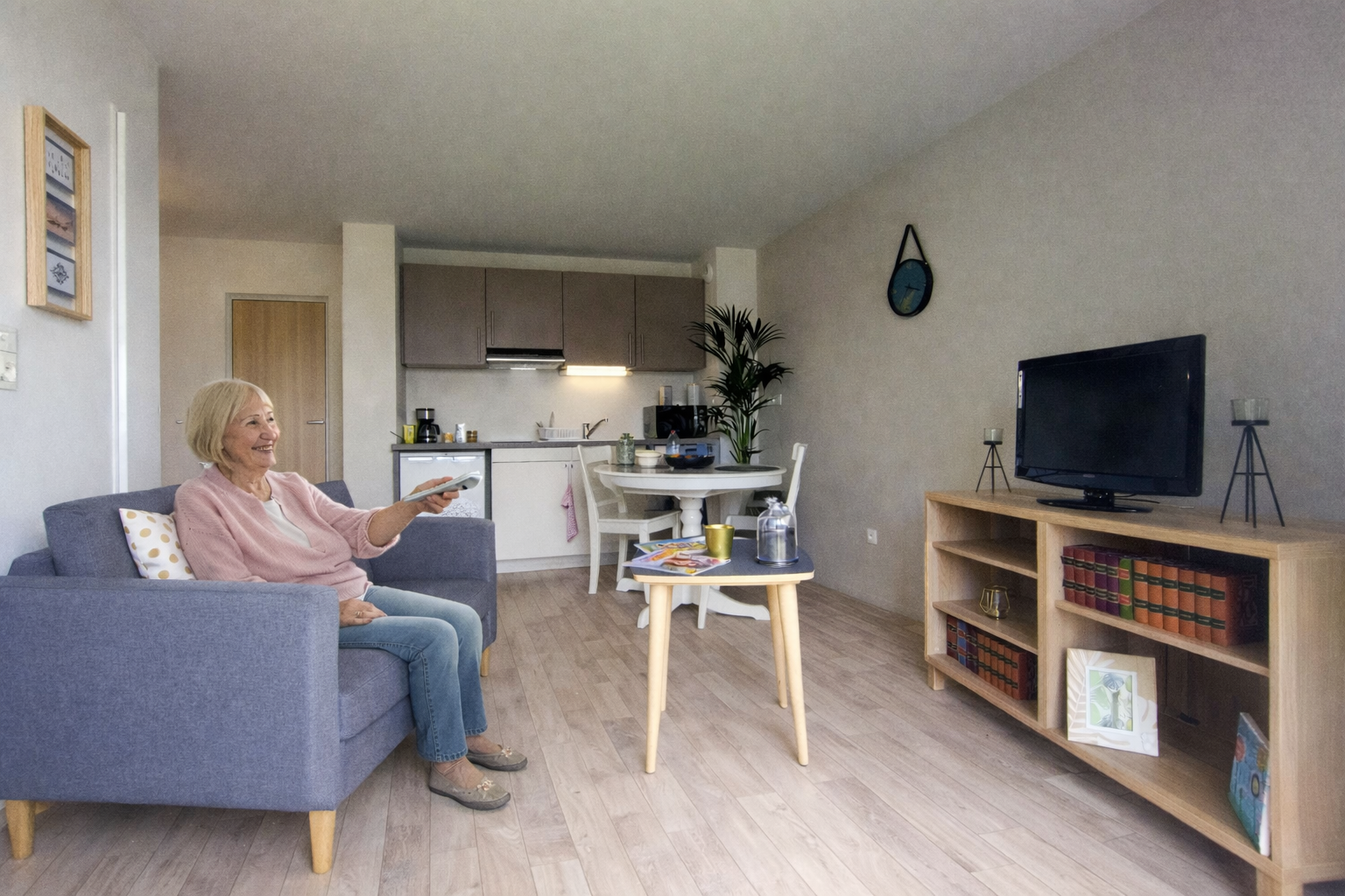 Dans un petit salon, une femme assise sur un canapé regarde la télévision, télécommande en main. On aperçoit la cuisine et la table à manger.