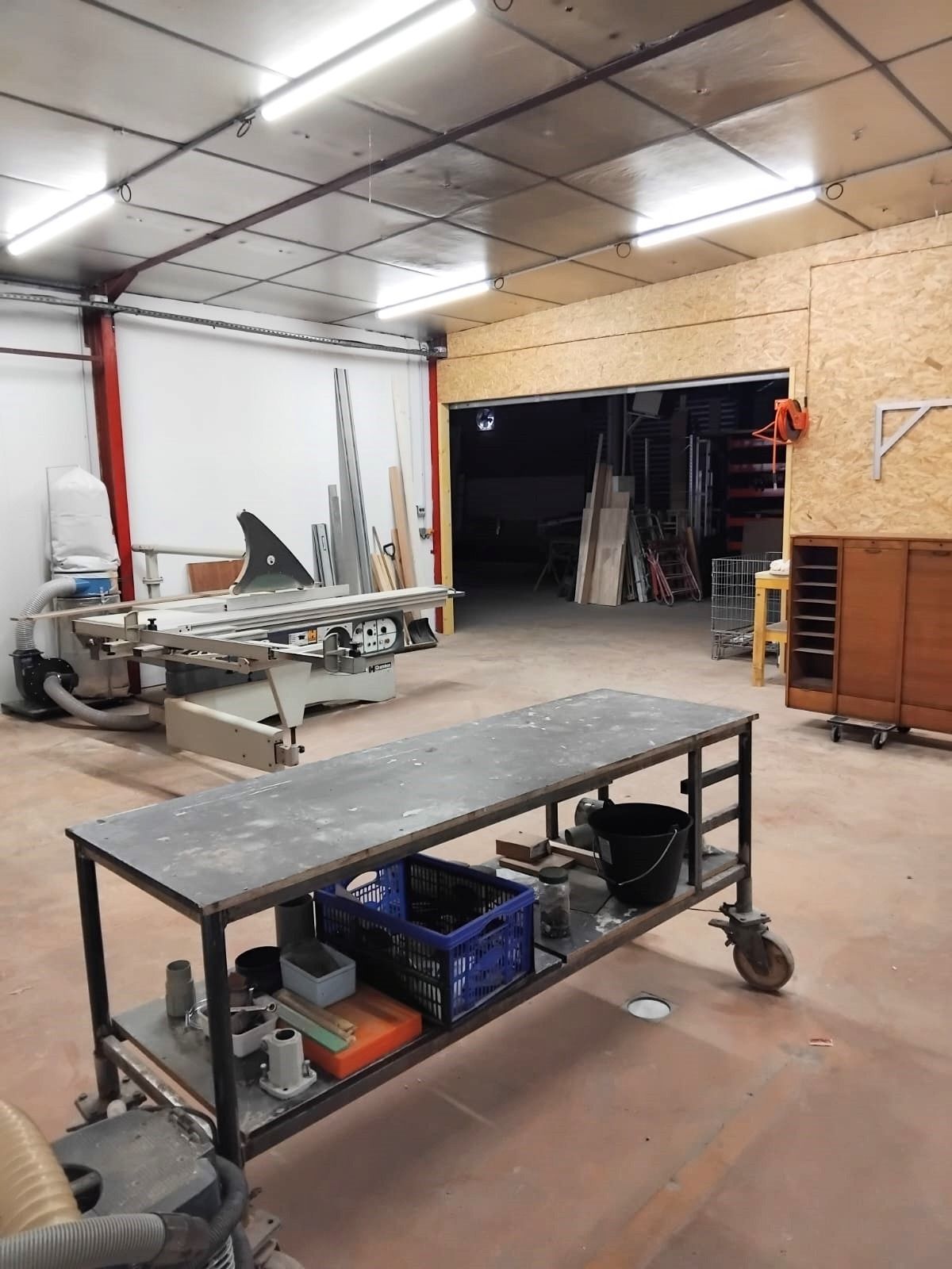 Intérieur d'atelier avec une table au premier plan, une scie et une porte ouverte menant au stockage.