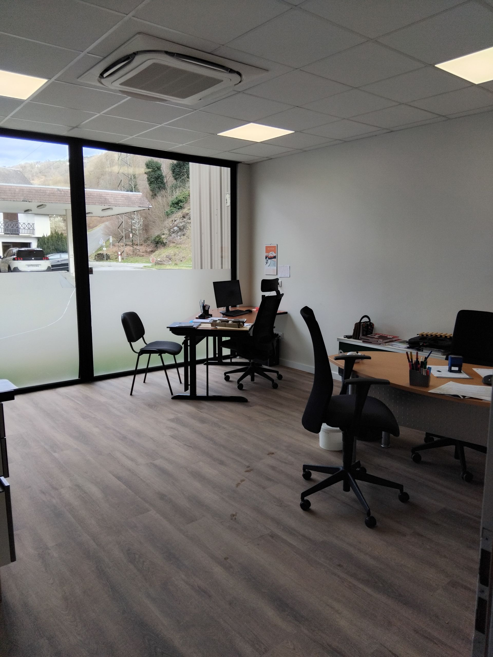 Intérieur de bureau avec bureaux, chaises, fenêtre et éclairage zénithal. Sol marron et vue extérieure d'un vert discret.