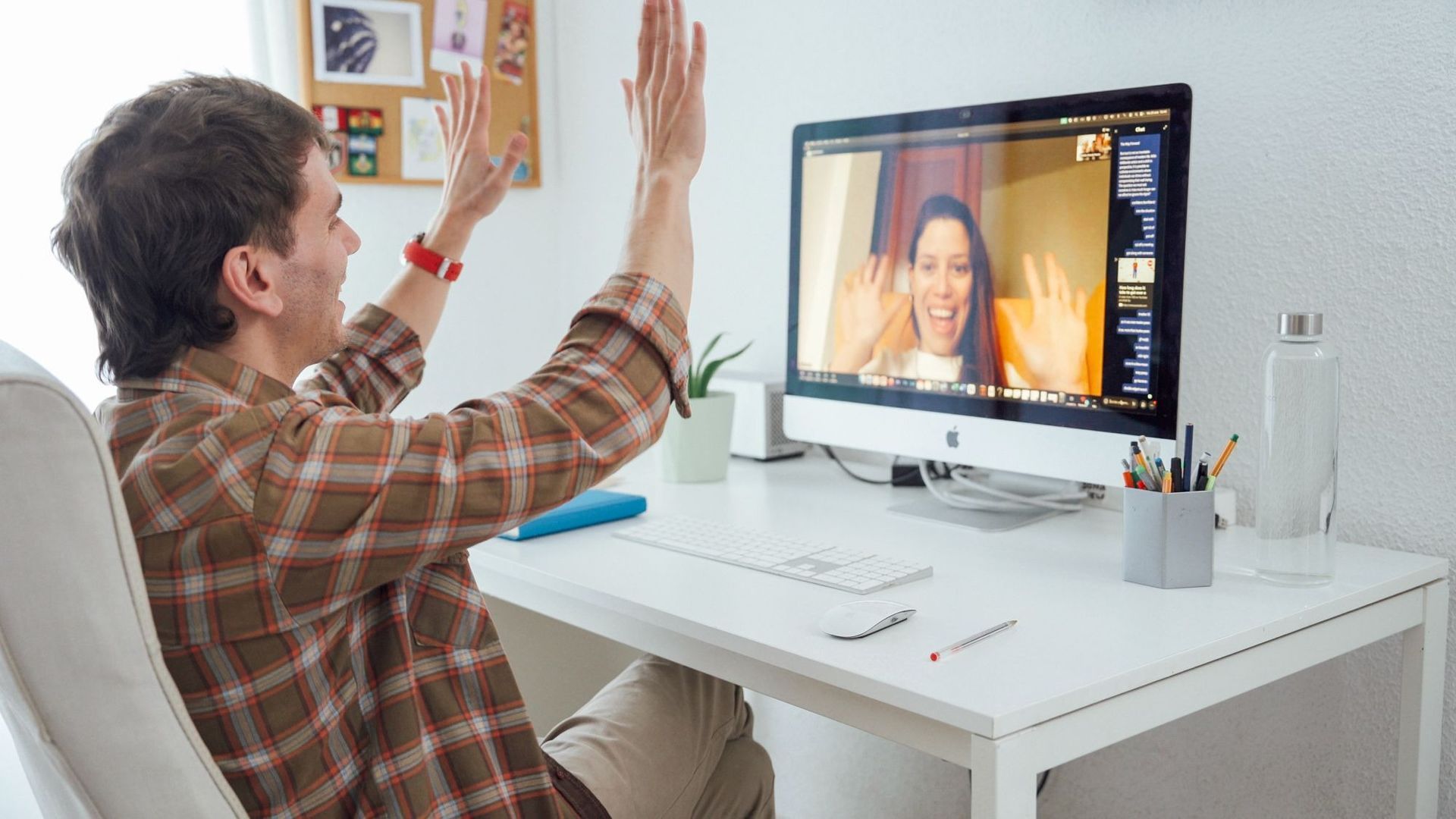 Una persona realiza una videollamada desde un escritorio blanco, saludando a un monitor en una luminosa oficina en casa.