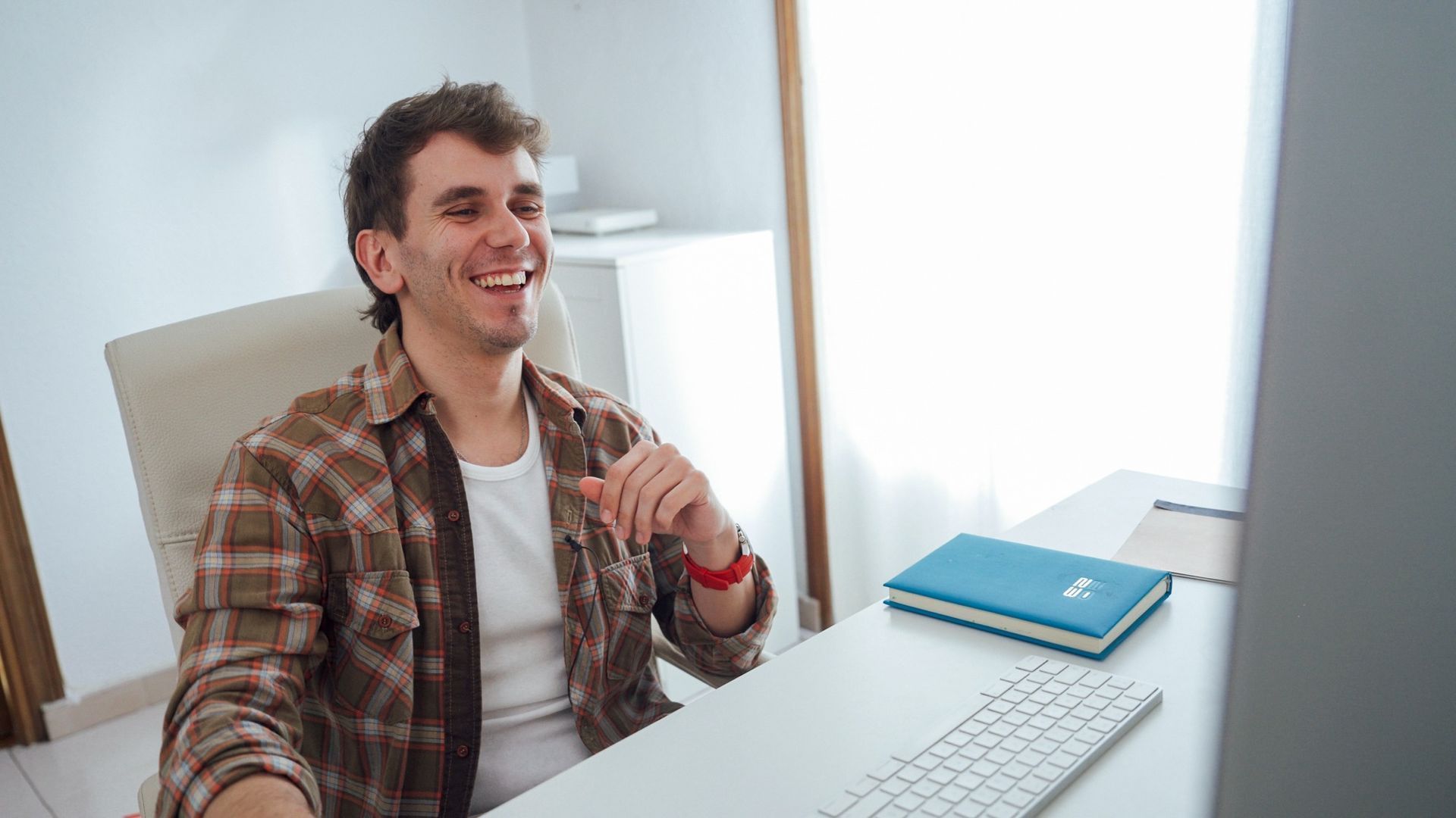Hombre sonriente con camisa a cuadros sentado en un escritorio, hablando en una videollamada