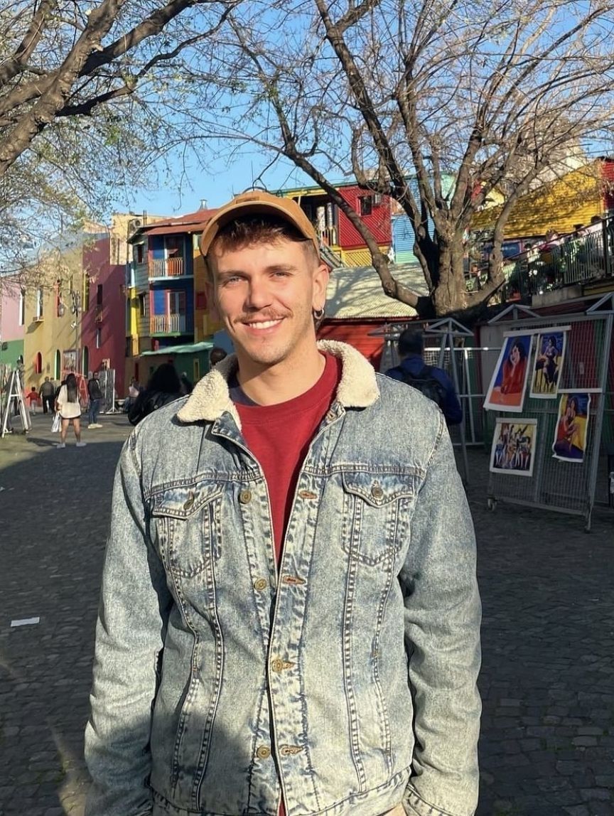Una persona sonriente con una chaqueta vaquera está de pie al aire libre frente a edificios coloridos y árboles desnudos.