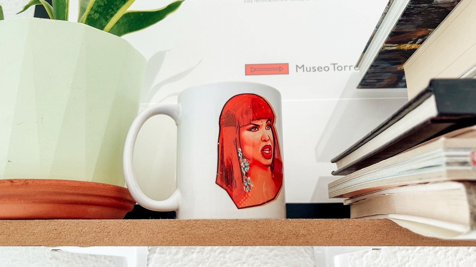 Taza roja con el retrato estilizado de una mujer sobre un escritorio junto a libros y una planta en maceta.