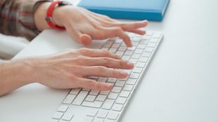 Manos tecleando en un teclado blanco junto a un cuaderno azul.