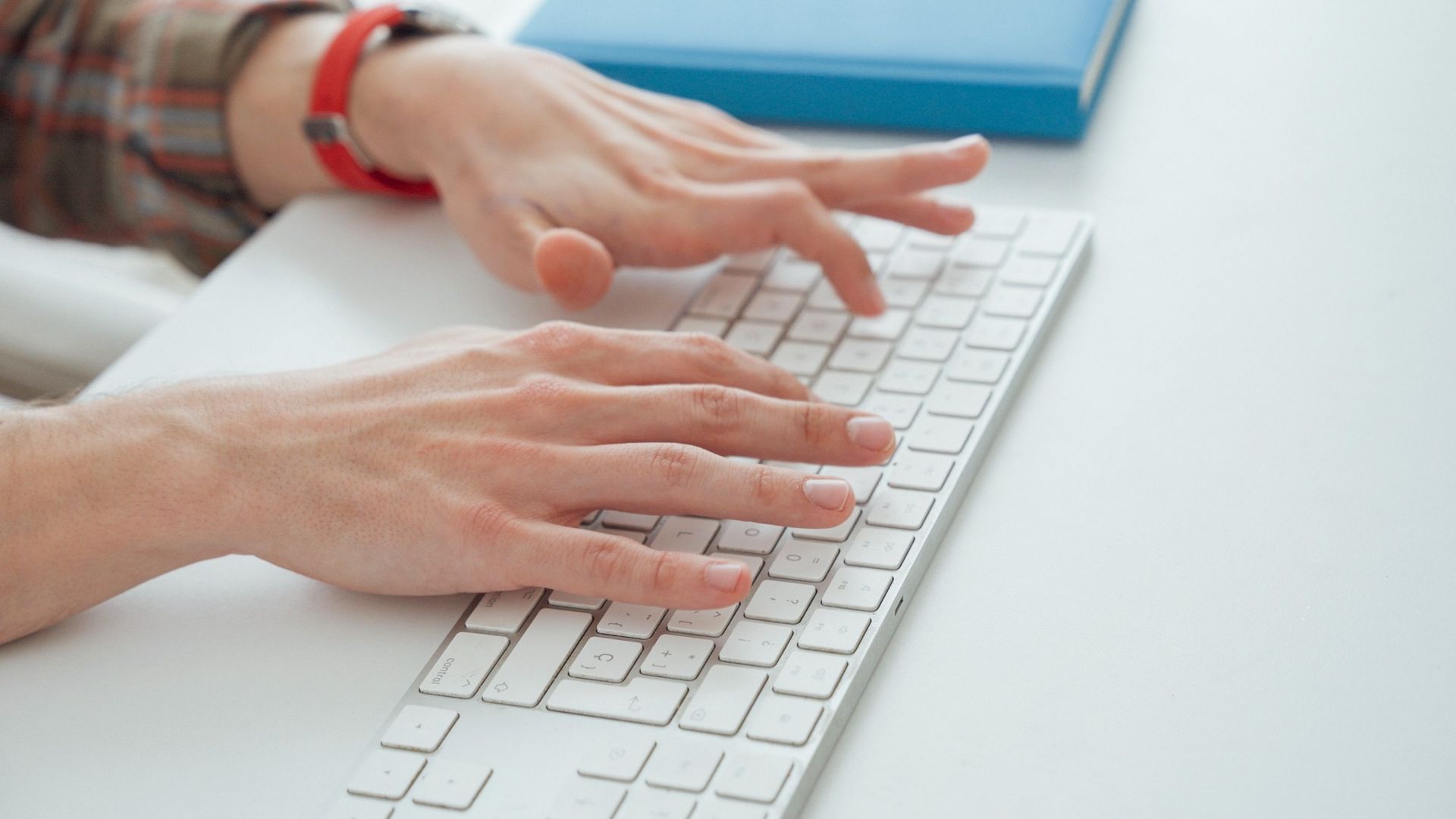 Manos tecleando en un teclado blanco junto a un cuaderno azul sobre un escritorio.