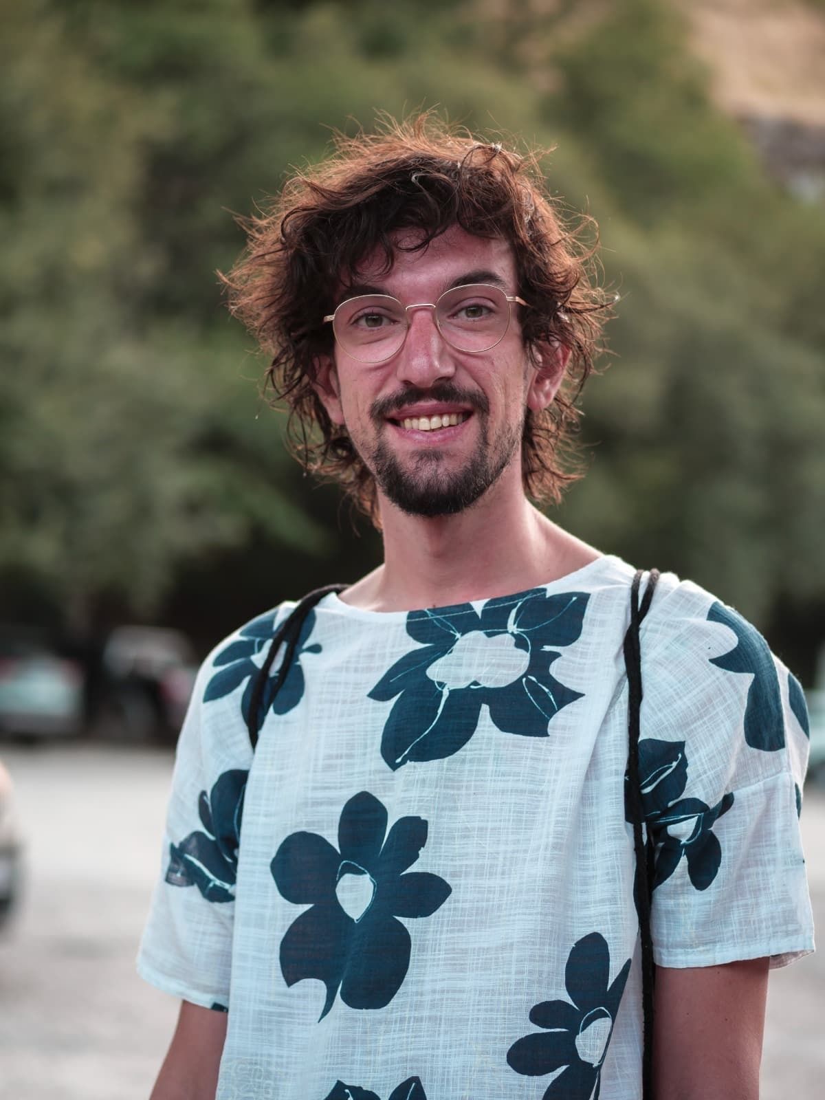 Persona sonriente con cabello rizado y gafas, vestida con una camisa blanca con flores azules, al aire libre.