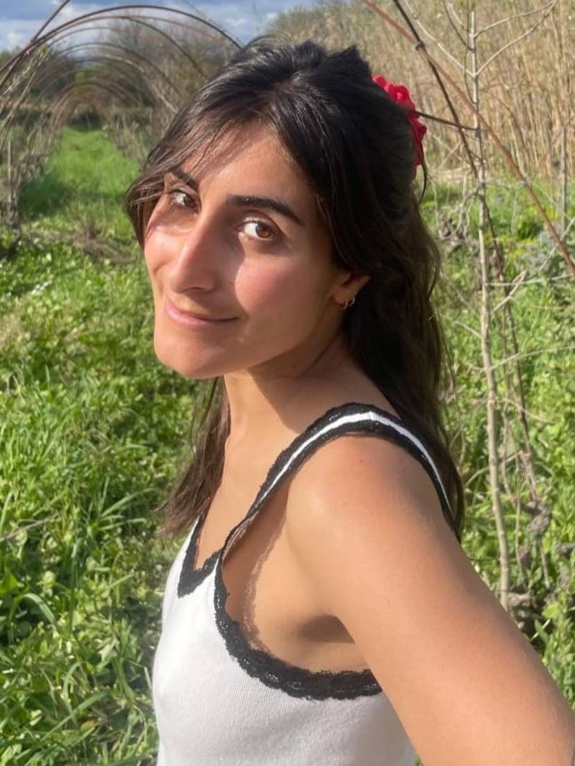 Mujer de pie en un campo de hierba, mirando a la cámara, con una camiseta blanca de tirantes con ribetes negros.