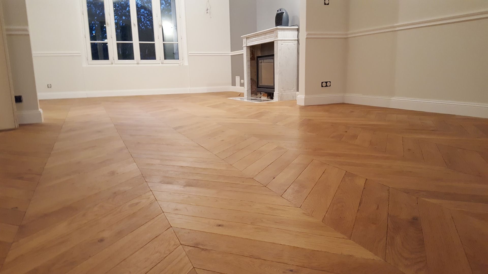Parquet à chevrons dans une pièce avec cheminée, fenêtre et murs blancs.
