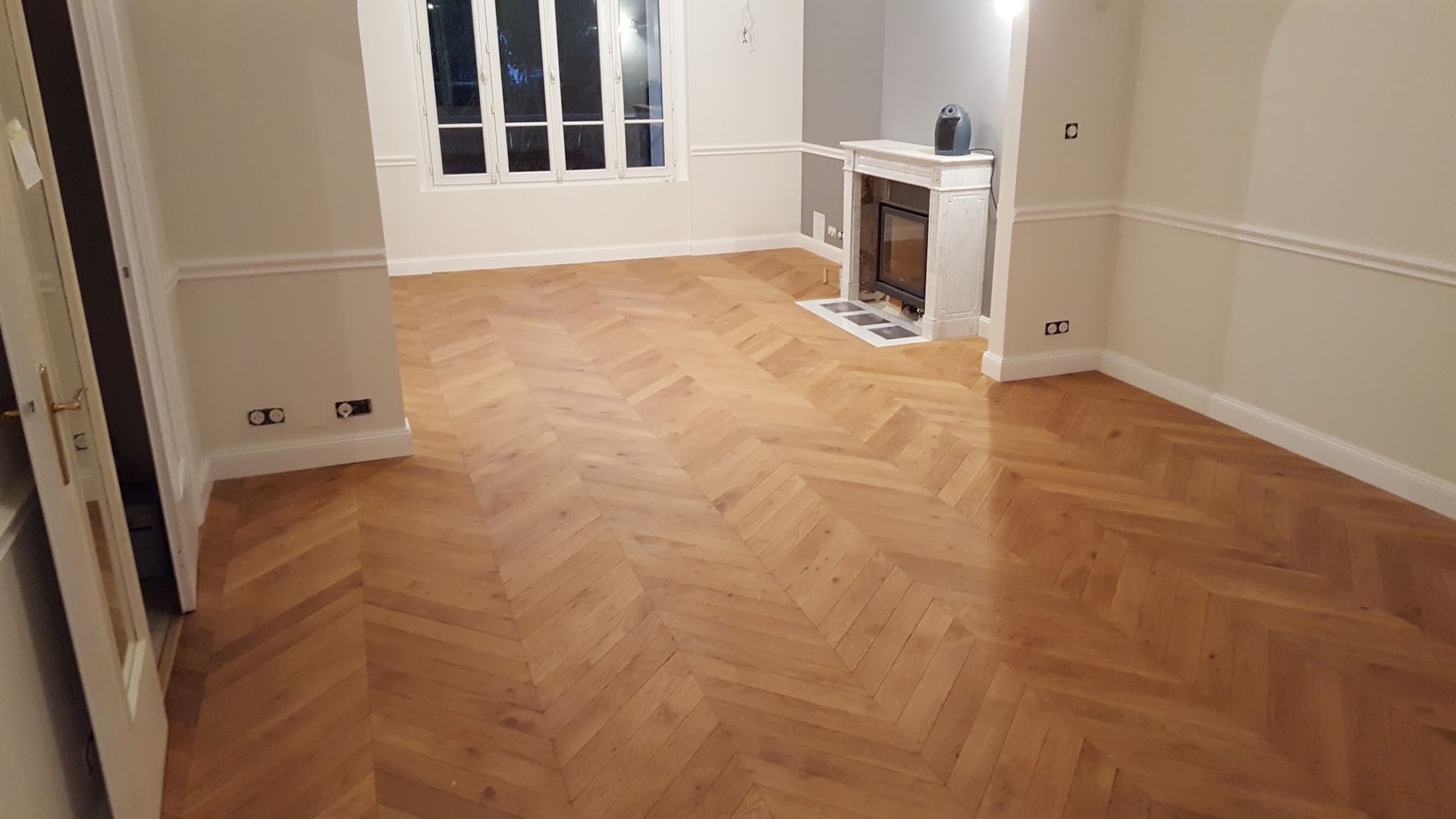 Pièce vide avec parquet, moulures blanches, fenêtre et cheminée.