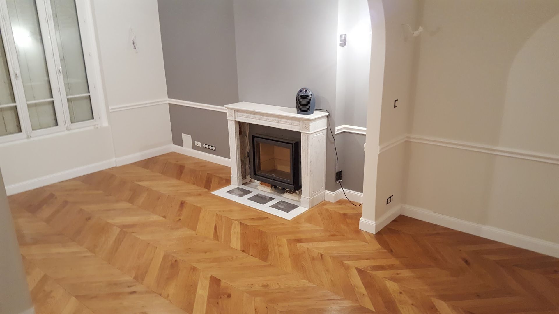 Pièce avec parquet, cheminée, fenêtre et murs gris/blancs.