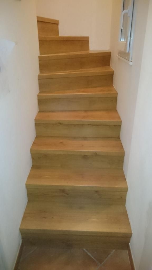 Escalier en bois montant, avec des marches claires et des murs blancs de chaque côté.