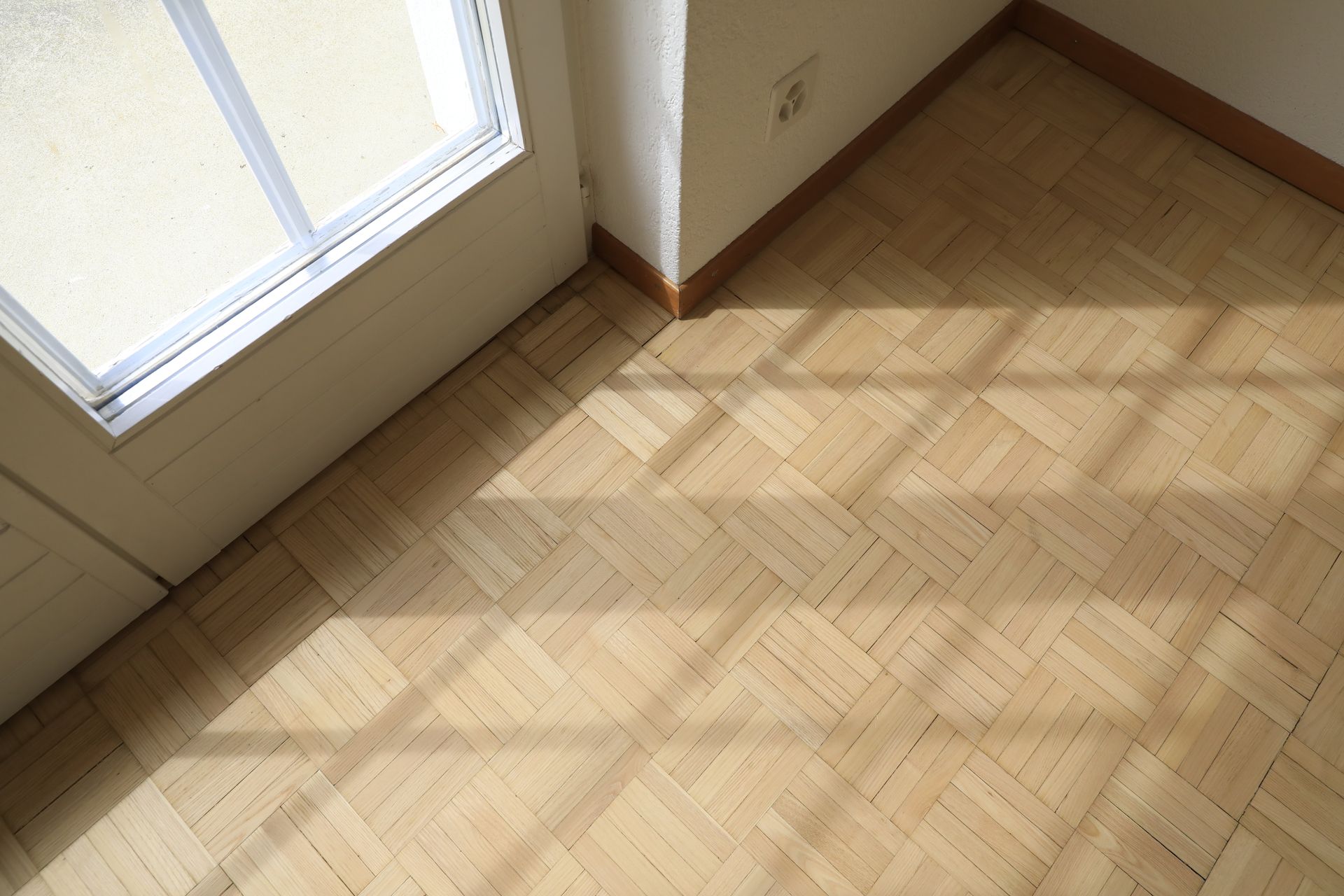 Parquet en bois avec des ombres projetées par une fenêtre, à côté d'un mur blanc et d'un cadre de fenêtre.