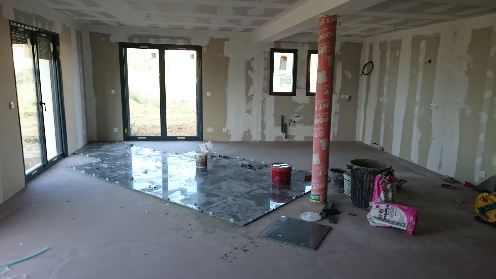 Pièce intérieure en travaux, avec cloisons sèches et poutre de soutien apparente. Un grand carrelage foncé recouvre le sol.