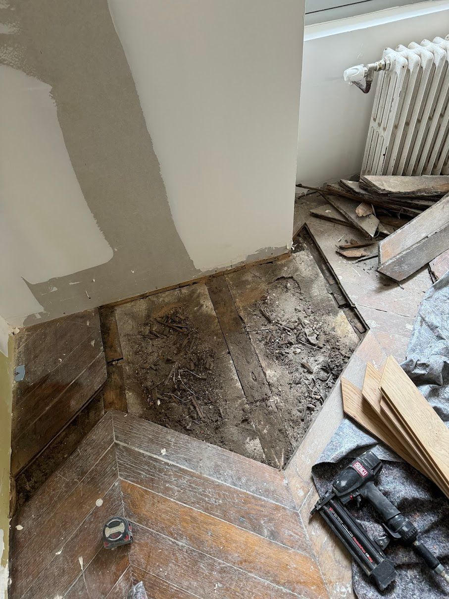 Plancher en mauvais état, laissant apparaître le sous-plancher. Cloueuse et débris au sol. Pièce avec un mur blanc et un radiateur.
