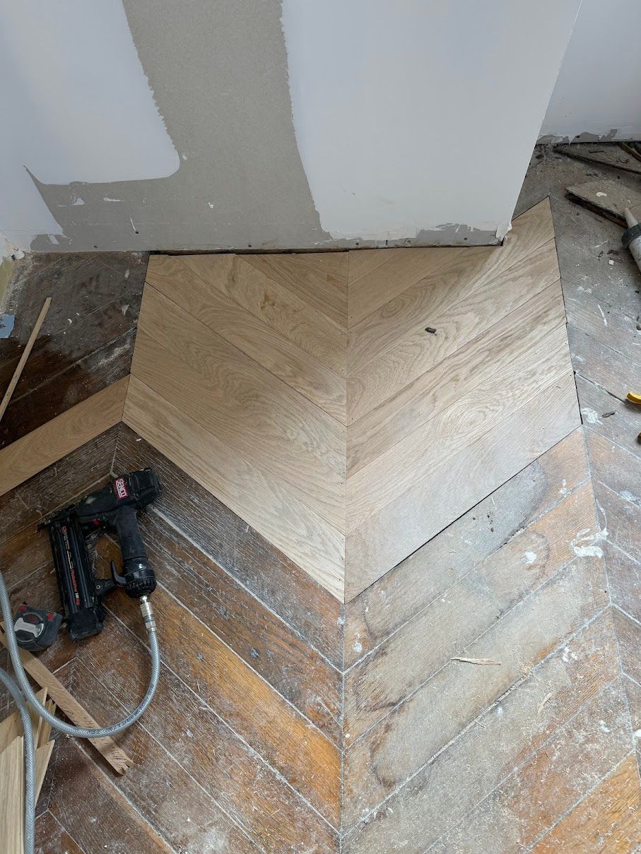 On installe un nouveau parquet clair à chevrons, à côté du parquet foncé existant. Un cloueur pneumatique se trouve à proximité.