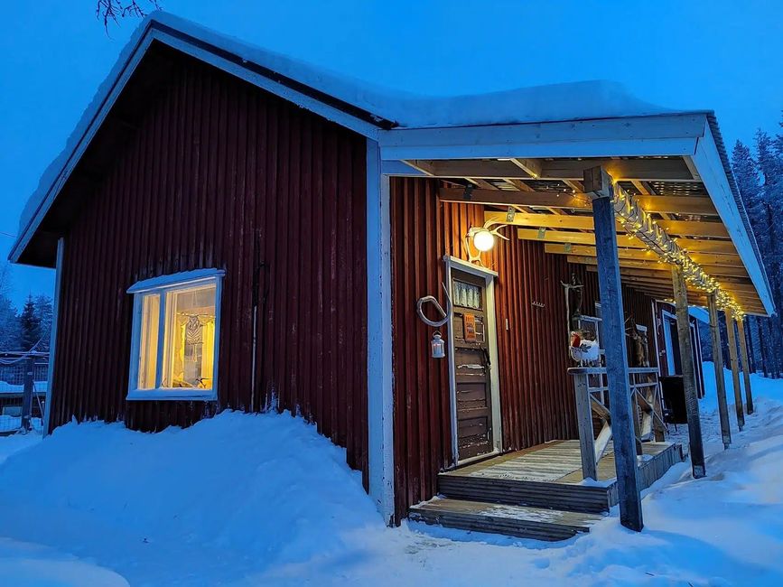 Lumisen peittämä punainen mökki, jossa on valaistu ikkuna ja kuisti sinisessä hämärässä.