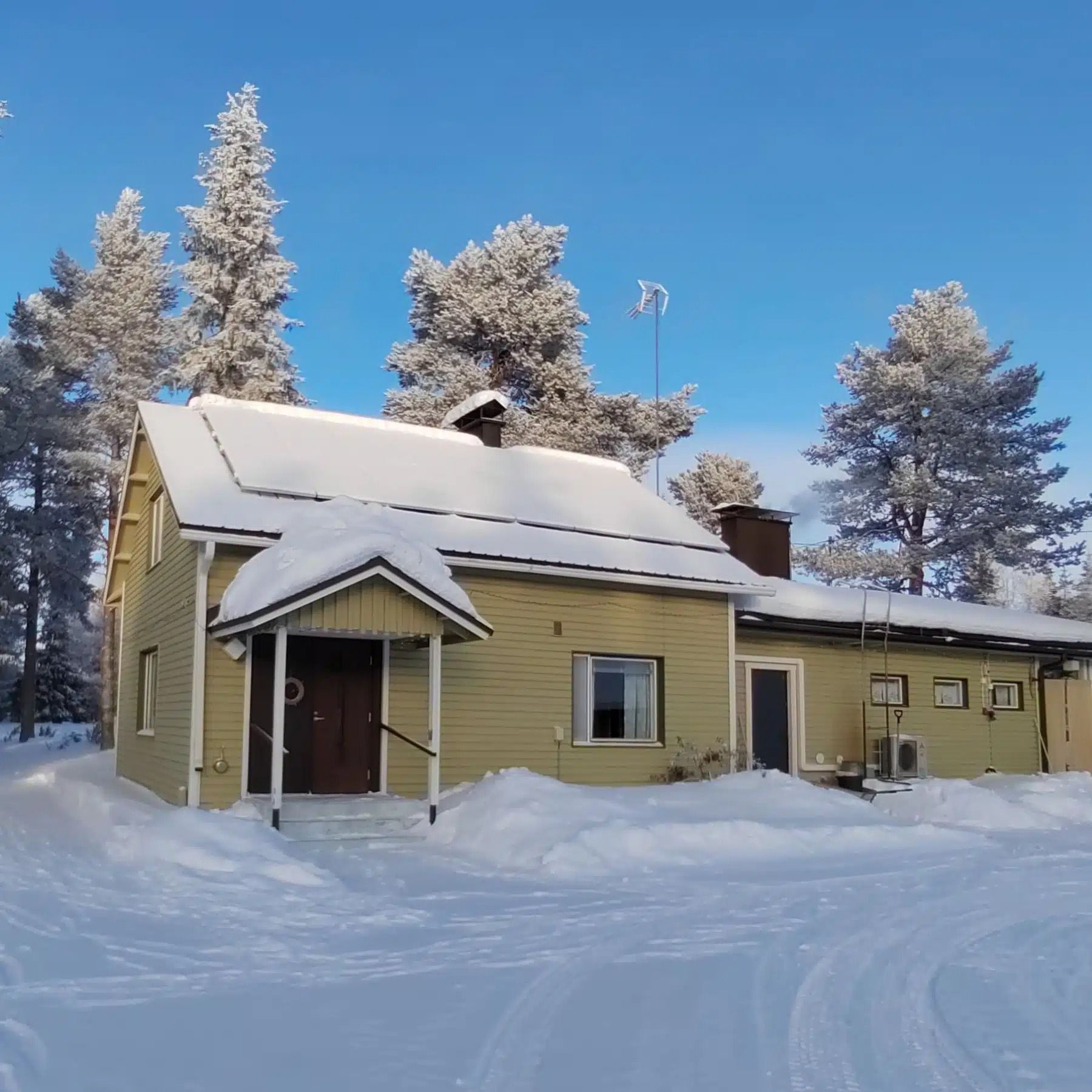 Lumen peittämä keltainen talo punaisella ovella ja huurteisilla puilla kirkkaan sinisen taivaan alla
