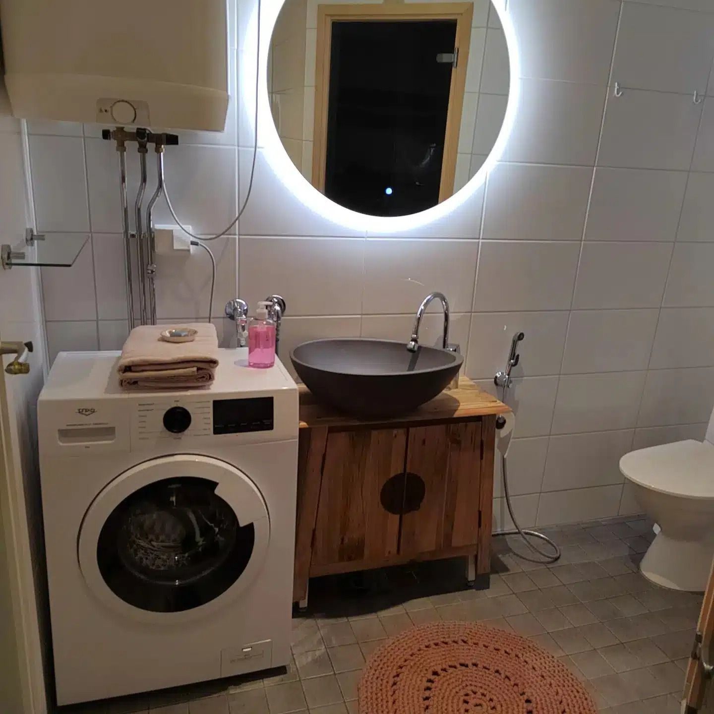 Kylpyhuone, jossa pesukone, puinen allaskaappi, pyöreä valaistu peili, laatoitettu seinät ja wc.