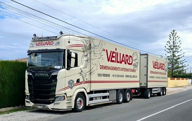 Camionnette avec remorque blanche stationnée sur une surface en béton, montagnes en arrière-plan. Le logo de l'entreprise est apposé sur le côté de la camionnette.