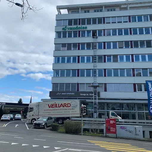 Un camion-grue décharge des meubles devant l'immeuble Vaudois. Des voitures circulent dans une rue sous un ciel bleu.
