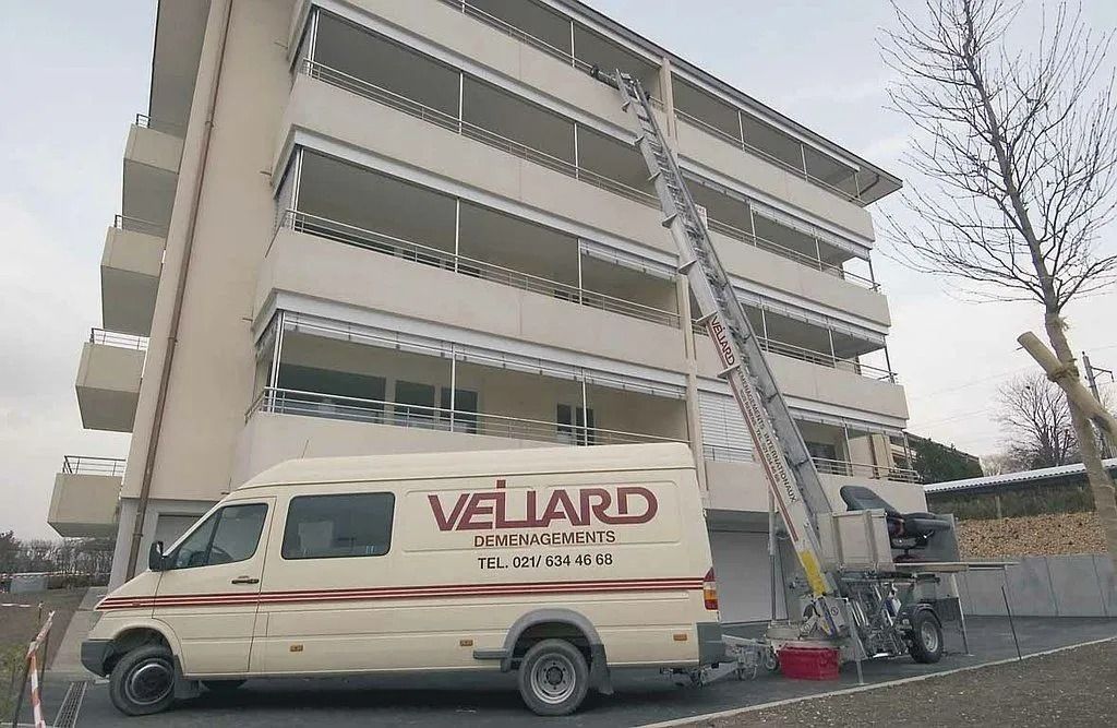 Camion de déménagement « Véliard » avec ascenseur, appuyé contre un immeuble de plusieurs étages, transférant des objets sur un balcon.