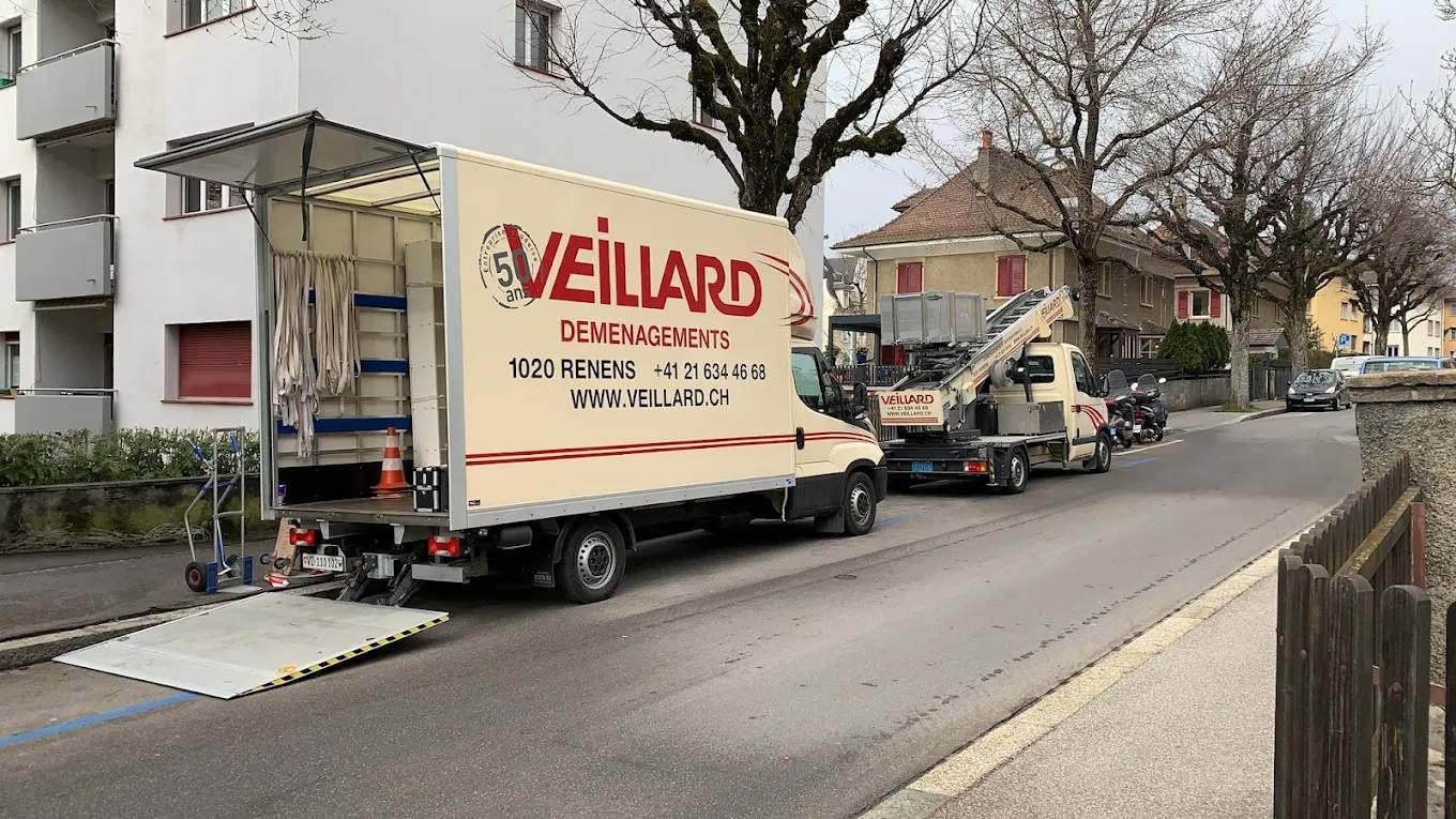 Un camion de déménagement Veillard à rampe ouverte est stationné dans une rue près d'un bâtiment, à côté d'un camion-grue.