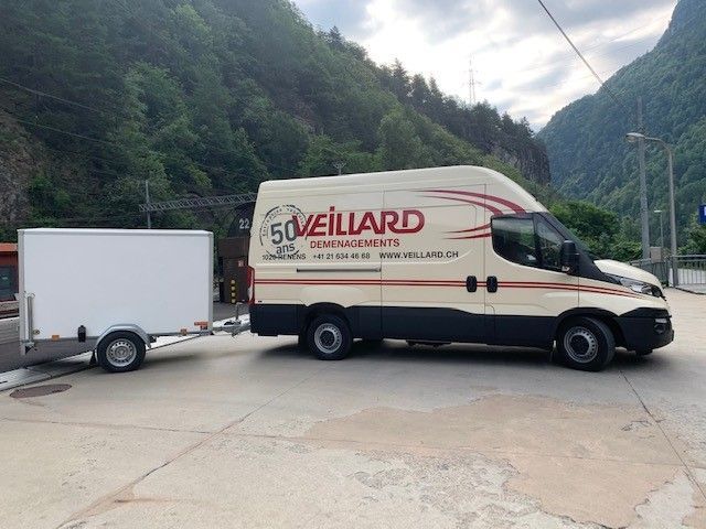 Camionnette avec remorque blanche stationnée sur une surface en béton, montagnes en arrière-plan. Le logo de l'entreprise est apposé sur le côté de la camionnette.