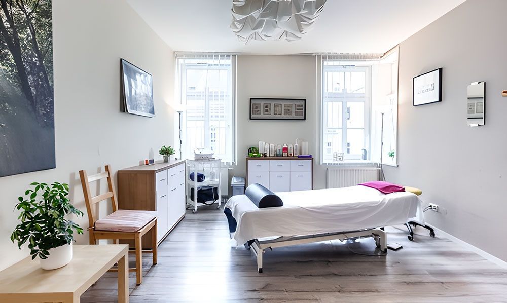 Massagetherapieraum mit Behandlungstisch, Fenstern und Kunstwerken an weißen Wänden.