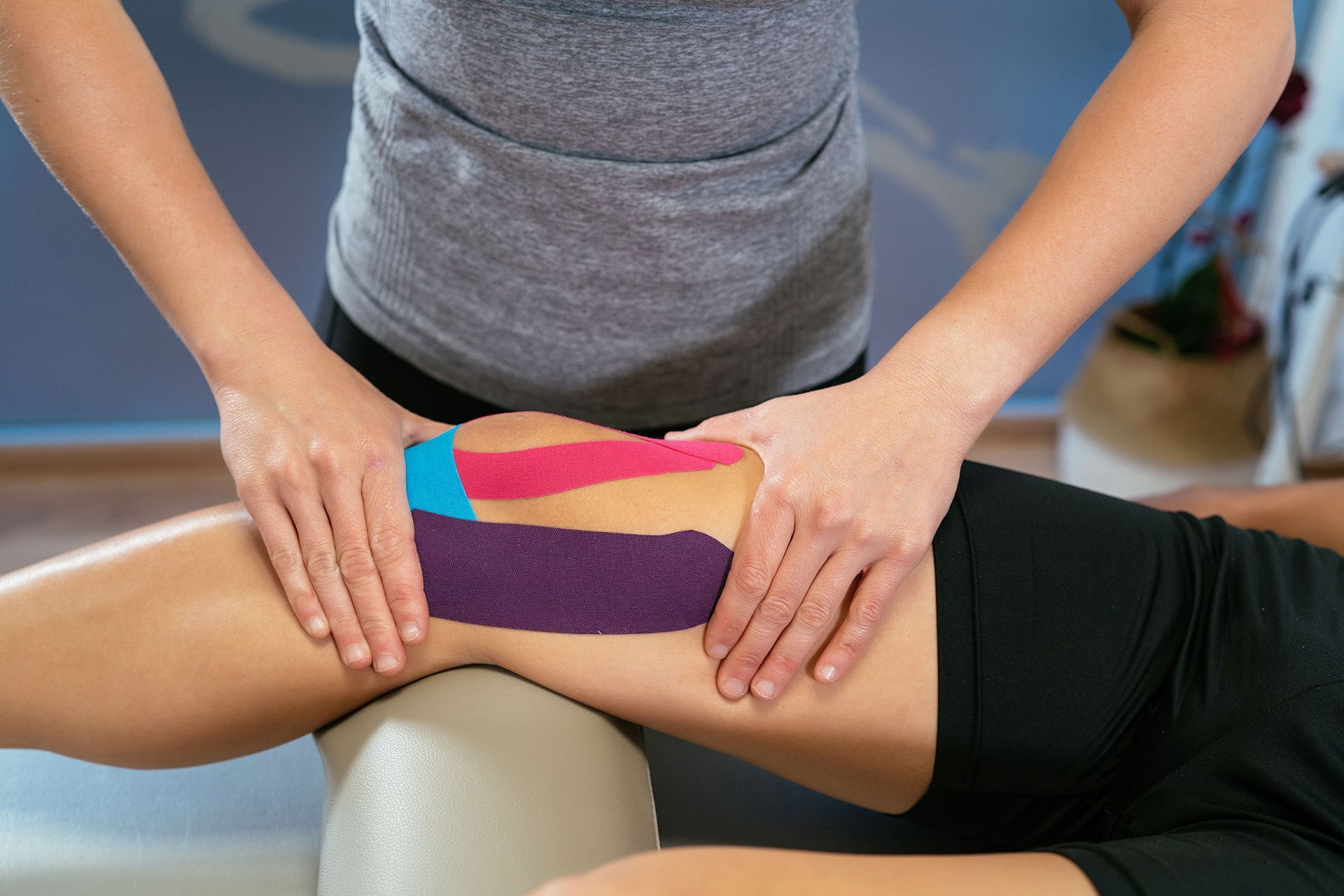 Das Bein einer Person mit buntem Kinesiologie-Tape wird von den Händen einer anderen Person beurteilt.