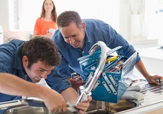 Dos personas con camisas azules trabajan en un fregadero de cocina con una caja de herramientas metálica cerca