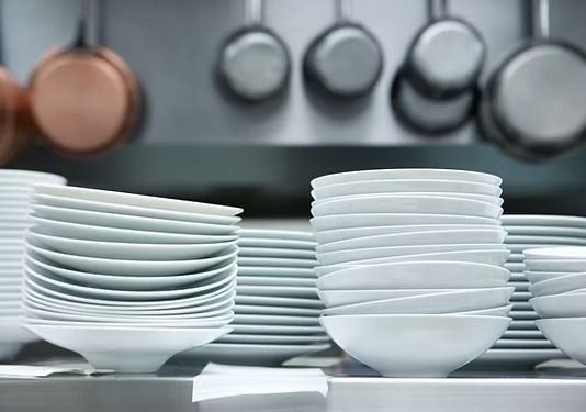 Montones de platos y cuencos blancos reposan sobre una encimera de cocina de acero inoxidable, con sartenes