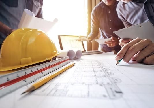 Un casco amarillo, una regla y un lápiz descansan sobre unos planos mientras unos profesionales discuten los planes al fondo.