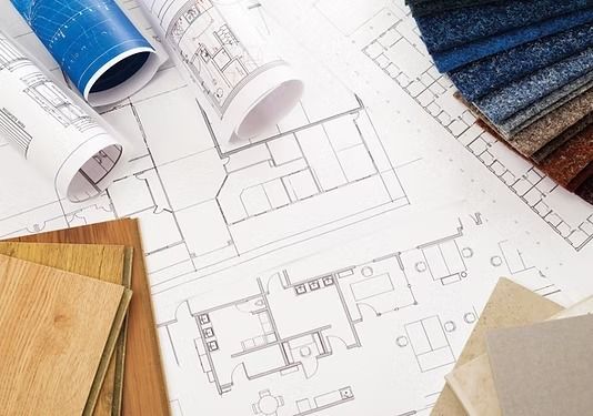 Planos arquitectónicos dispuestos con láminas enrolladas, tablones de madera para el suelo y muestras de moqueta azul.